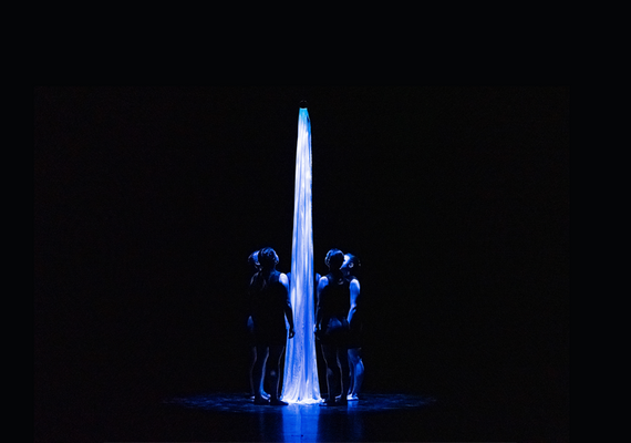 Four dancers surrounding a white cloth. They are all lit in blue light, in front of a black background.